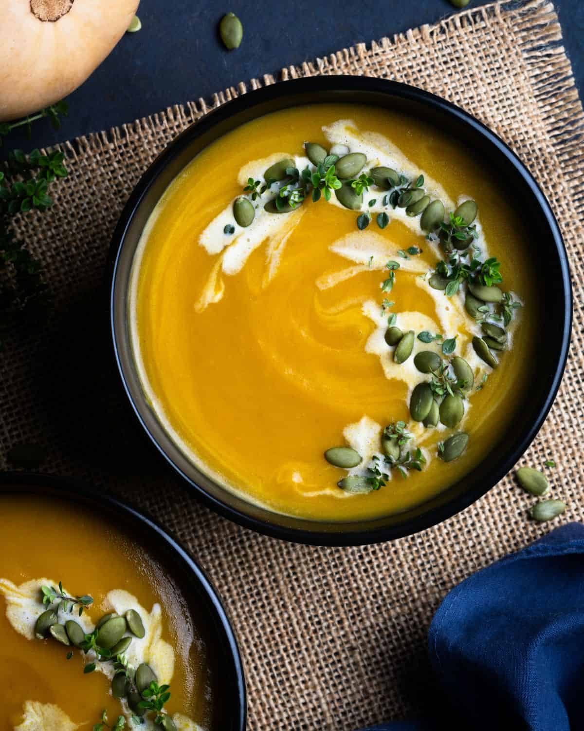A bowl of creamy Butternut Squash Bisque soup is garnished with a swirl of cream, green pumpkin seeds, and fresh herbs, sitting on a piece of burlap cloth. Another bowl of soup is partially visible beside it.