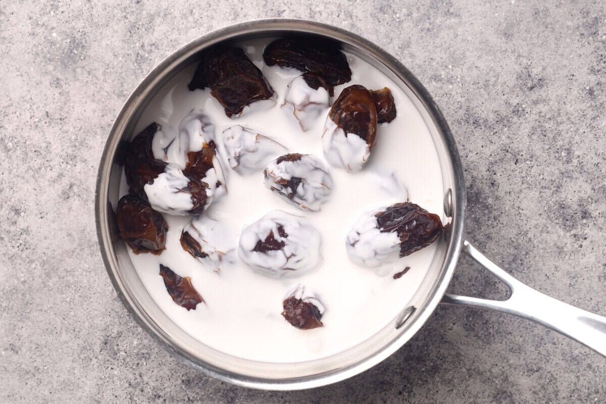 A metal saucepan filled with dates and coconut milk sits on a gray textured surface. The dates are partially submerged and coated in the coconut milk, creating a contrasting dark and white appearance.