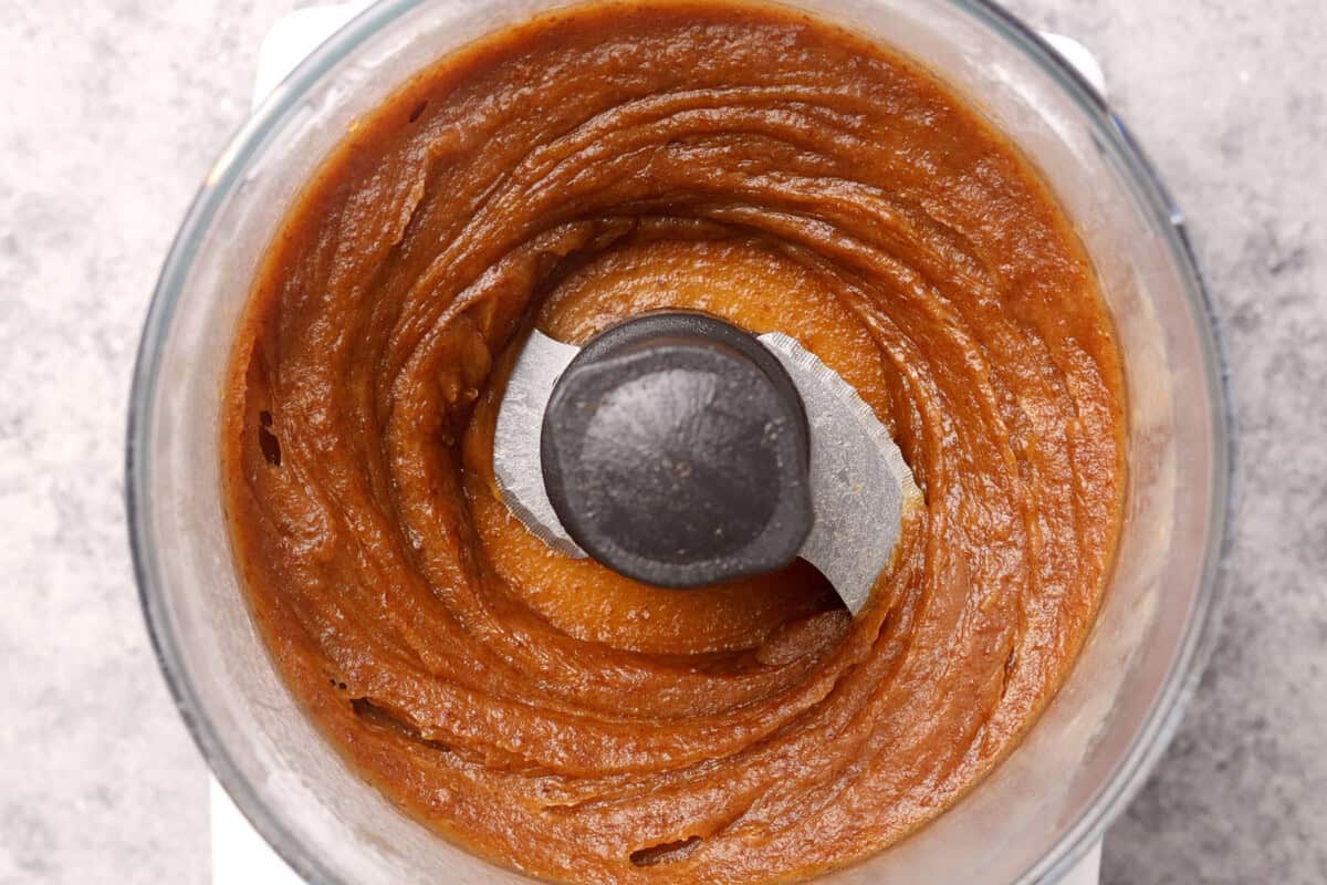 A top-down view of a food processor containing smooth, brown date caramel with a creamy texture spiraled around the metal blade.