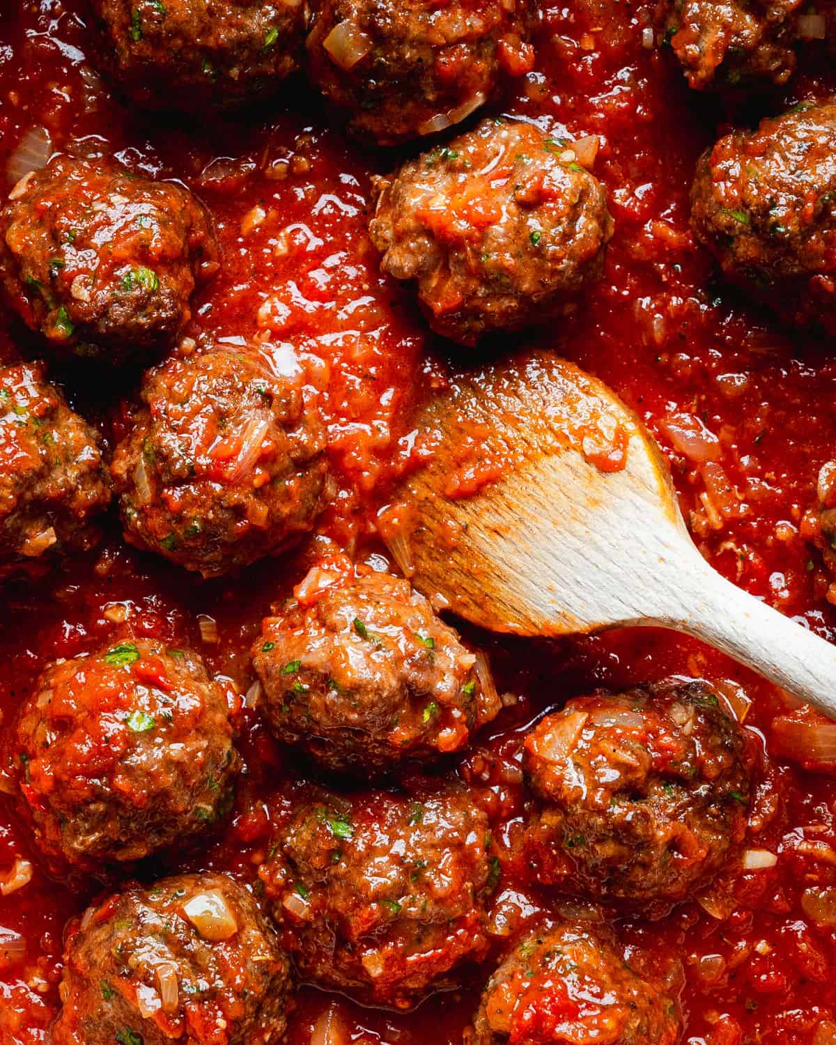 Mexican meatballs simmering in rich red chipotle sauce, garnished with herbs. A wooden spoon rests among the meatballs, stirring the sauce.