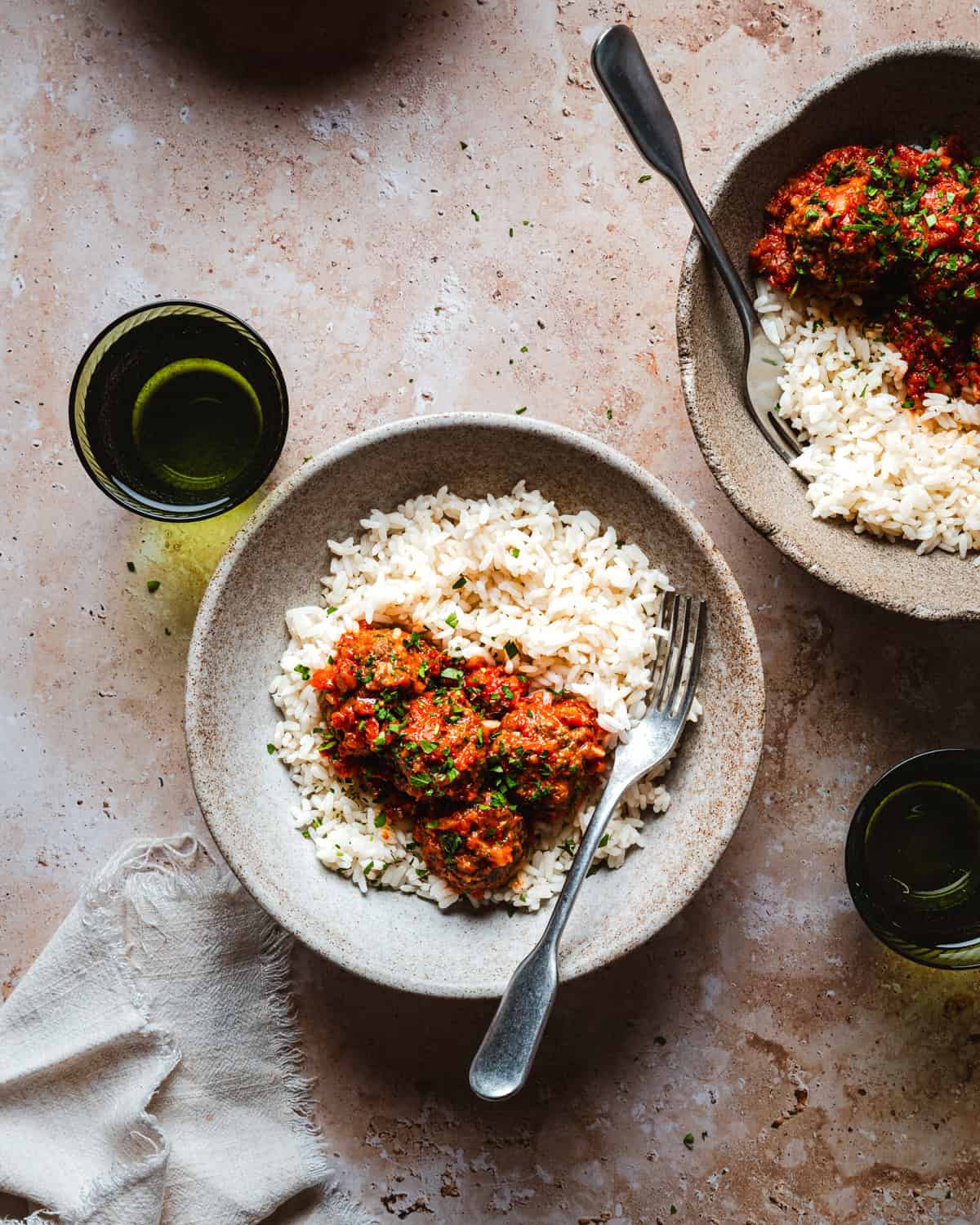 Two ceramic bowls filled with white rice topped with Mexican meatballs in chipotle sauce and herbs, each with a fork. Two green glasses and a beige napkin are also on the light brown textured table.