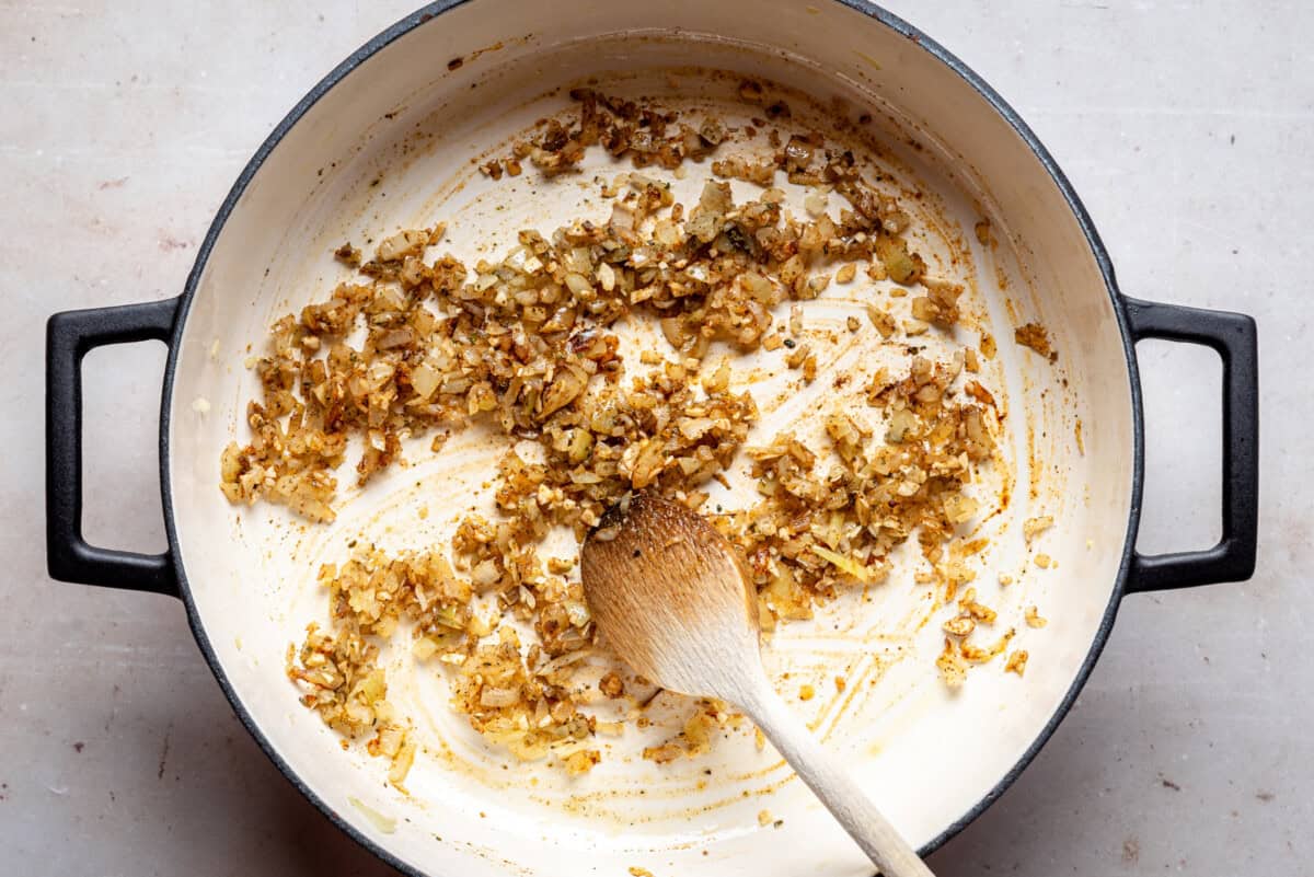 Chopped onions sautéing in a large white pot with black handles, some browned bits scattered, and a wooden spoon resting inside.