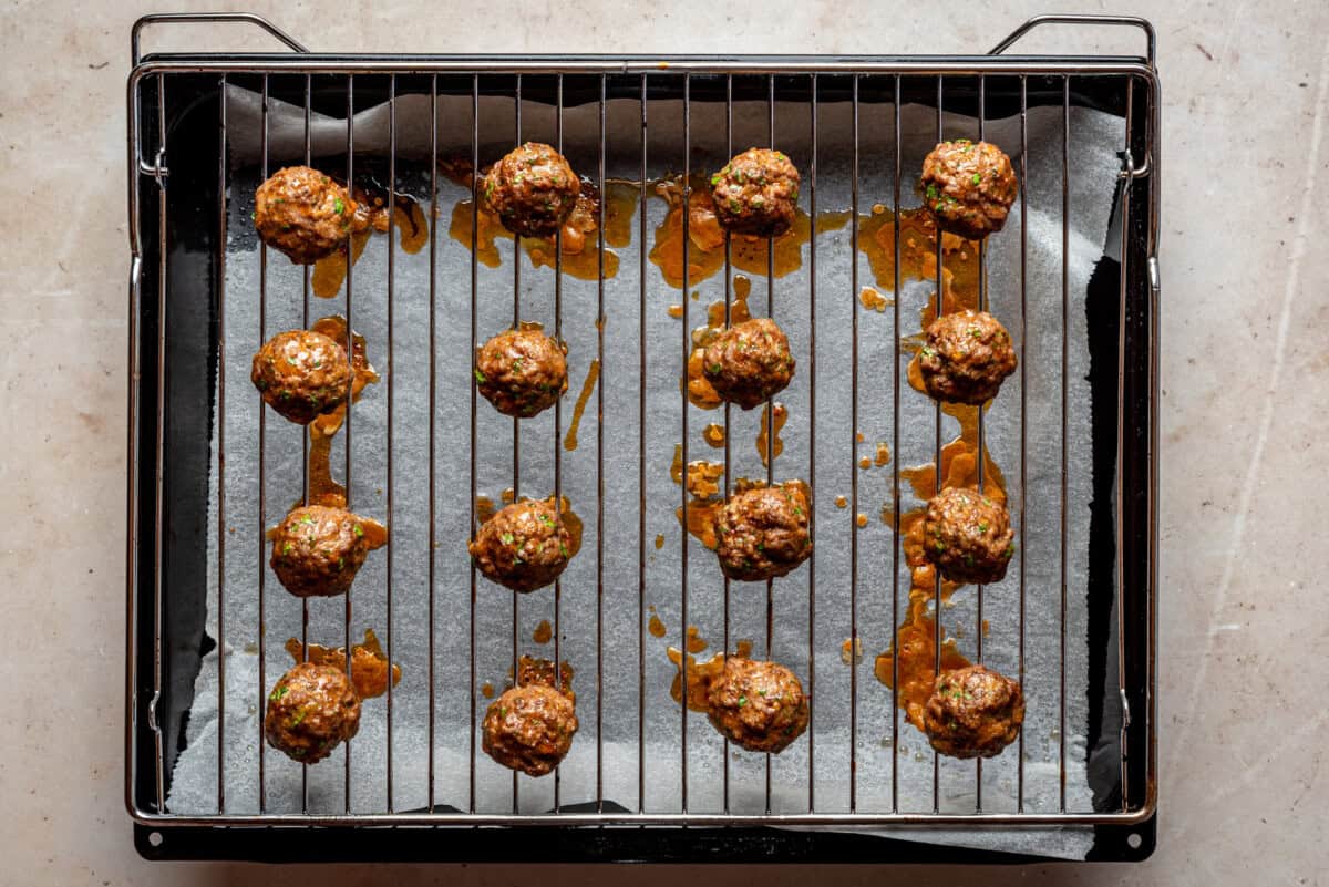 A baking tray lined with parchment paper holds a metal rack with evenly spaced cooked meatballs, with some oil drips visible on the paper below.