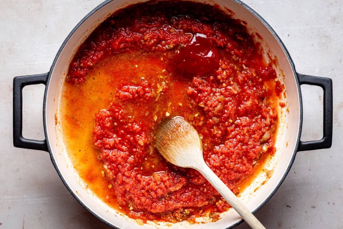 A pot filled with simmering tomato chipotle sauce, showing a mix of red tomatoes and oil, with a wooden spoon resting inside. The pot sits on a light-colored surface.