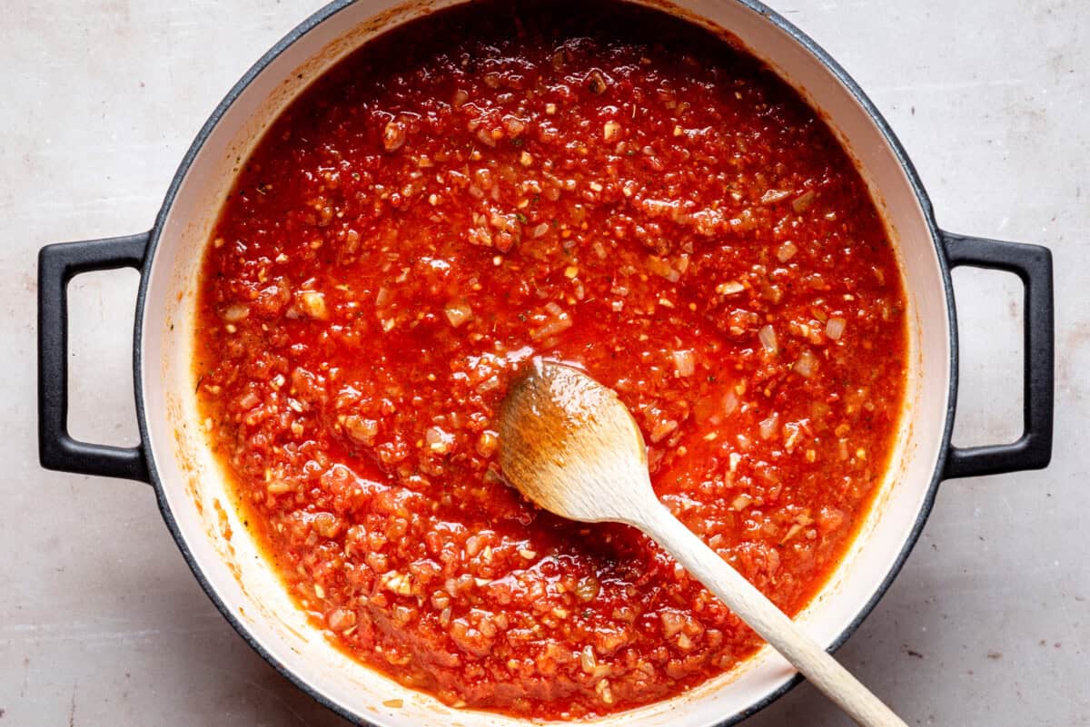 A pot of chunky red chipotle sauce with visible pieces of onion and possibly meat, stirred with a wooden spoon, sits on a light-colored surface.