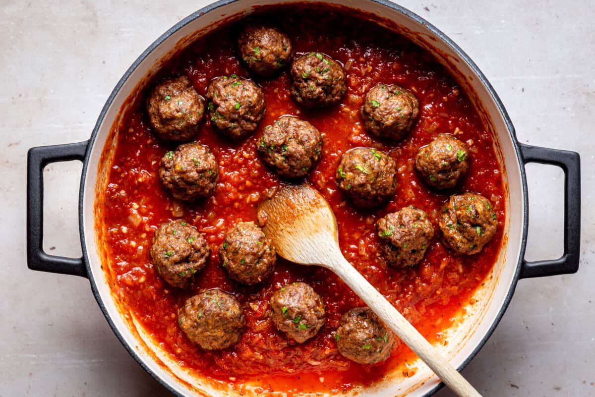 A pot of Mexican meatballs in tomato sauce with a wooden spoon resting inside, seen from above. The meatballs are arranged on the sauce in a round, black-handled pot.