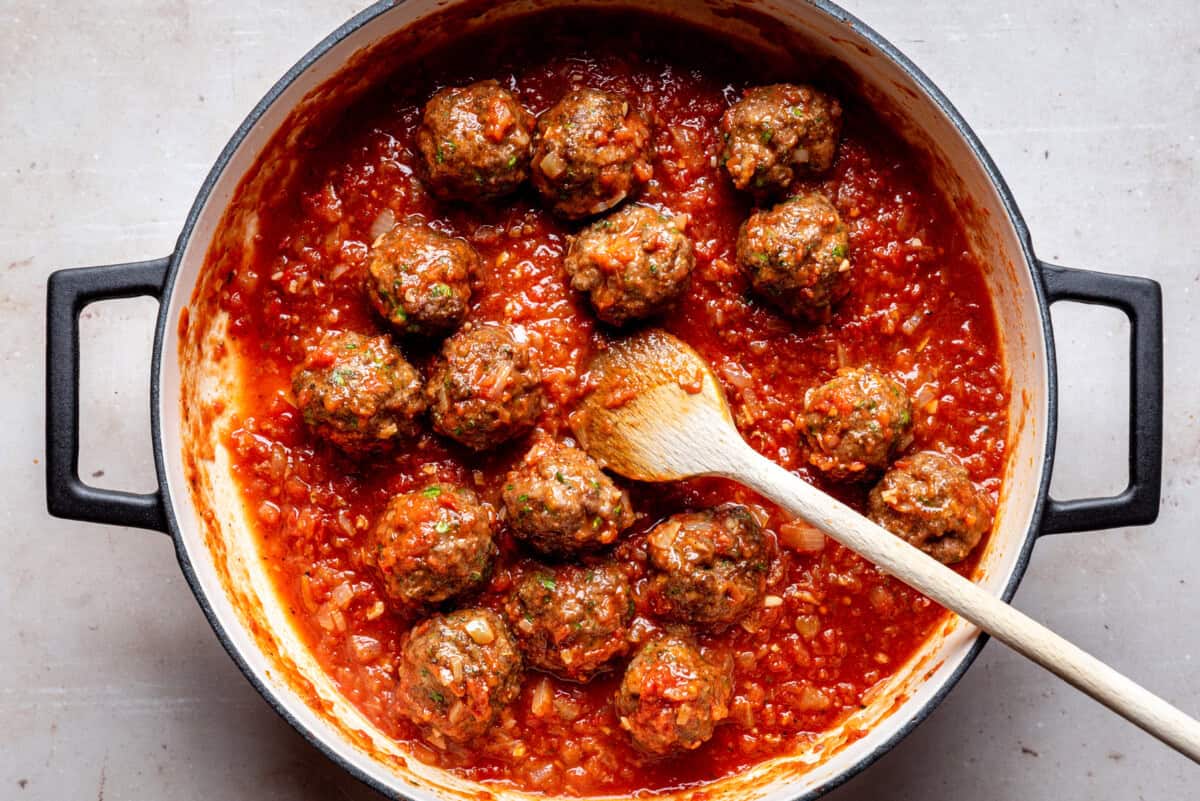 A pot of Mexican meatballs simmering in rich tomato sauce, with a wooden spoon resting inside, on a light-colored surface.