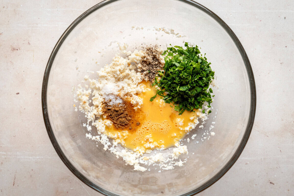 A glass bowl containing panko breadcrumbs, chopped parsley, beaten eggs, ground spices, salt, and pepper on a light-colored surface.