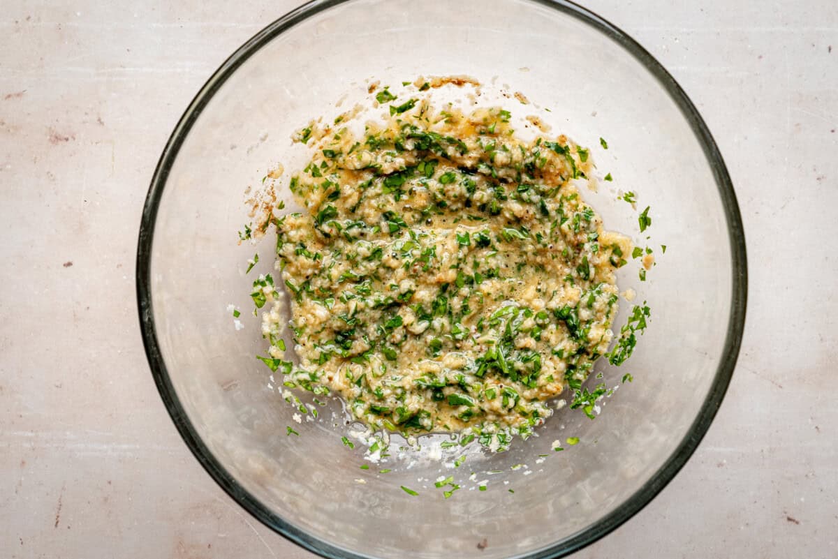 A glass bowl containing a mixed green and yellow herb paste or sauce, with visible chopped greens and a creamy, textured consistency, placed on a light-colored surface.