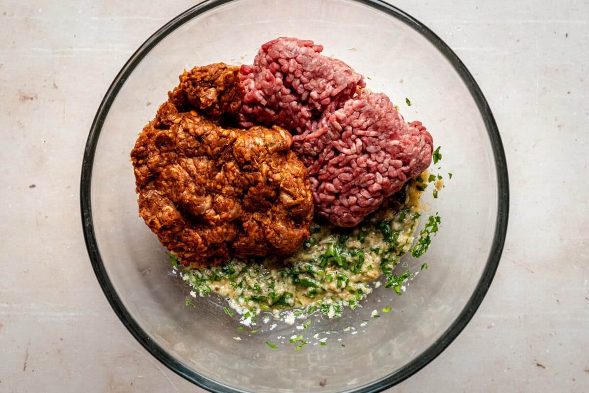 A glass bowl containing raw ground beef, raw Mexican chorizo, chopped herbs, and a mixture of wet ingredients on a light countertop.