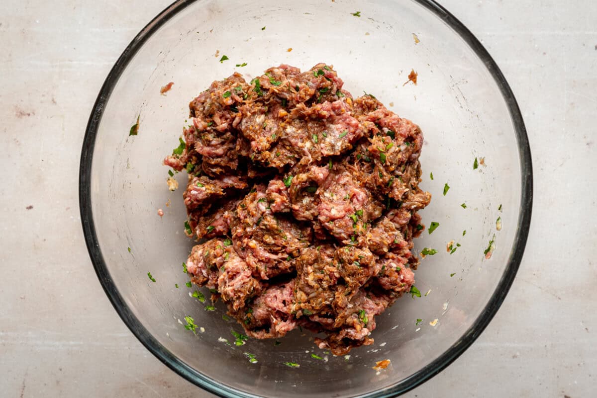 A clear glass bowl containing seasoned raw ground meat mixed with chopped herbs and spices, placed on a light-colored countertop.
