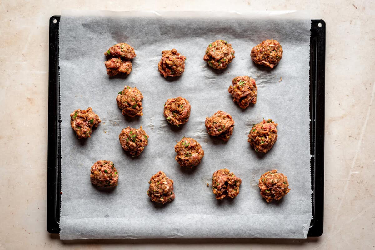 A baking tray lined with parchment paper holds 16 raw meatballs evenly spaced, ready to be cooked. The surface beneath the tray is a light, beige countertop.