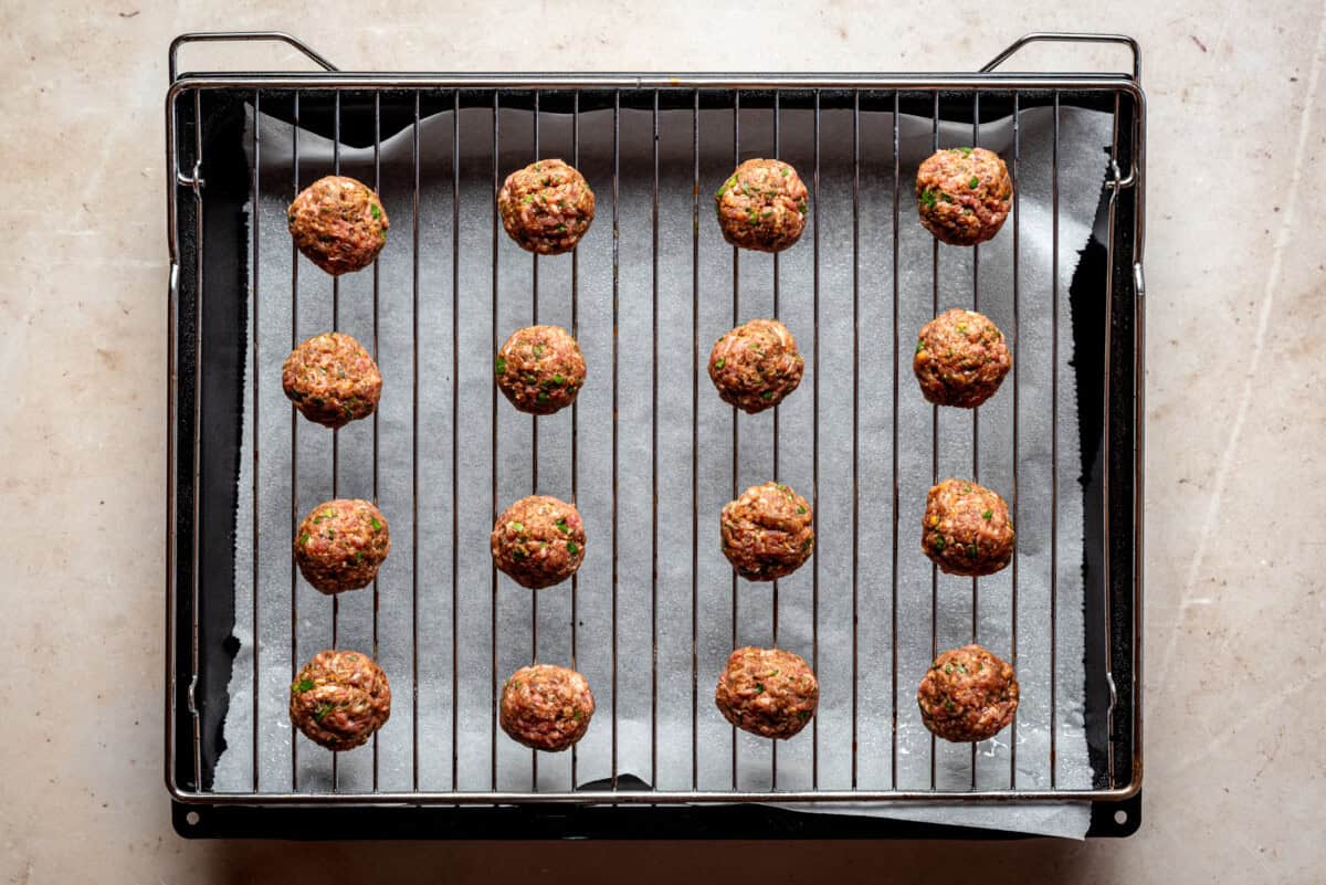 Raw meatballs arranged in rows on a wire rack set over a baking tray lined with parchment paper, ready for cooking.