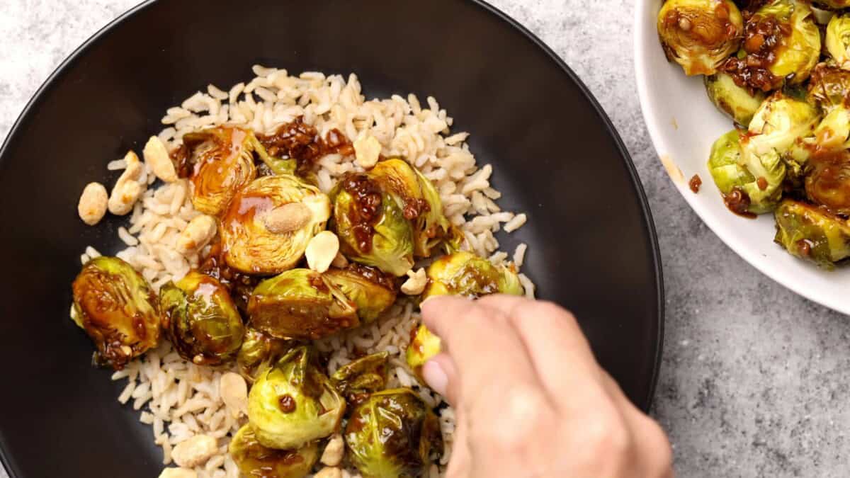 A hand sprinkles peanuts over a bowl of brown rice topped with roasted kung pao Brussels sprouts with a glossy sauce. A small bowl of extra Brussels sprouts sits nearby on a light countertop.