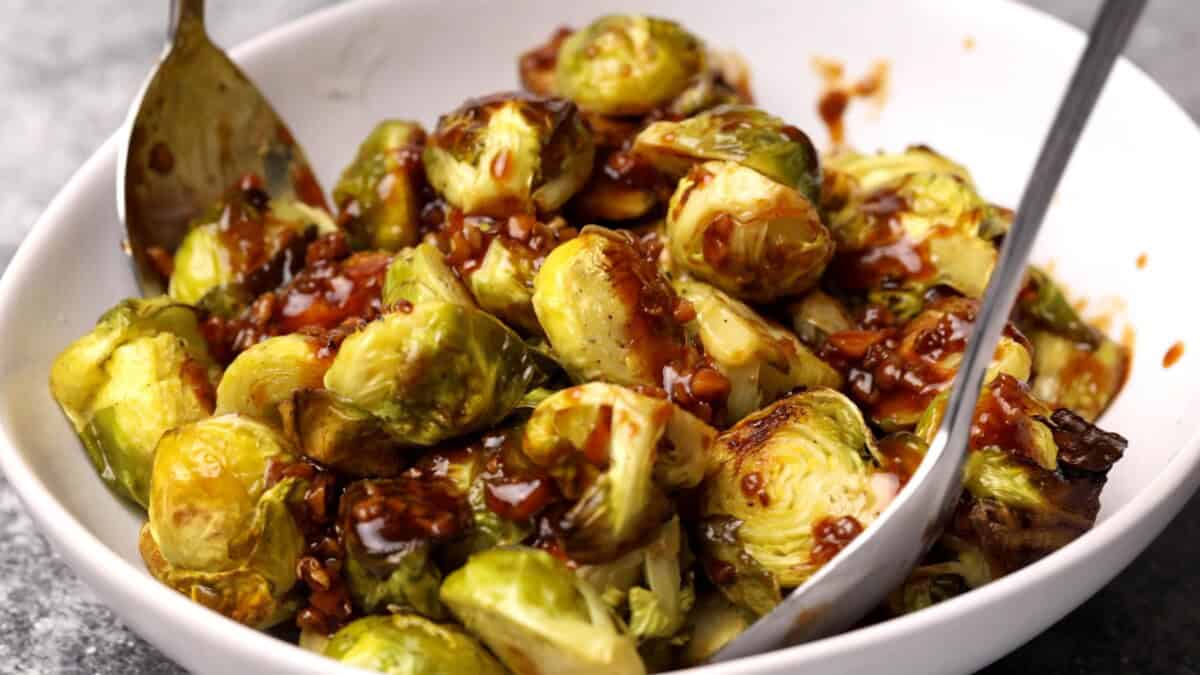 A bowl of roasted Brussels sprouts coated in a glossy kung pao sauce, with a spoon and fork resting in the bowl.