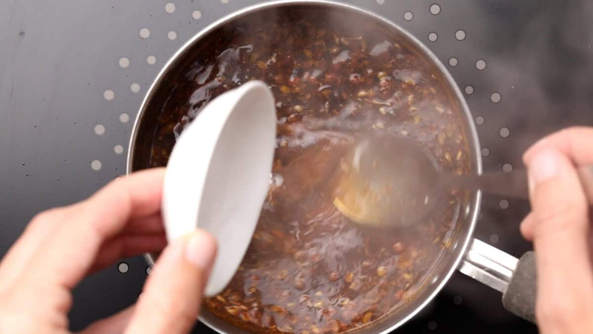 A hand holds a small white bowl of cornstarch slurry, pouring its contents into a pot of steaming, kung pao sauce with visible steam and spices while stirring the mixture on a stovetop.
