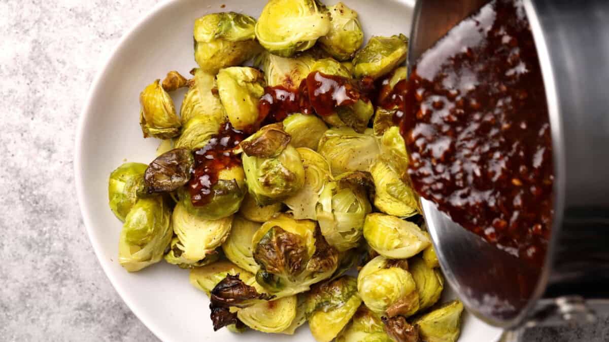 A white bowl filled with roasted Brussels sprouts, with a pot pouring kung pao sauce over the vegetables. The background is a light, textured surface.