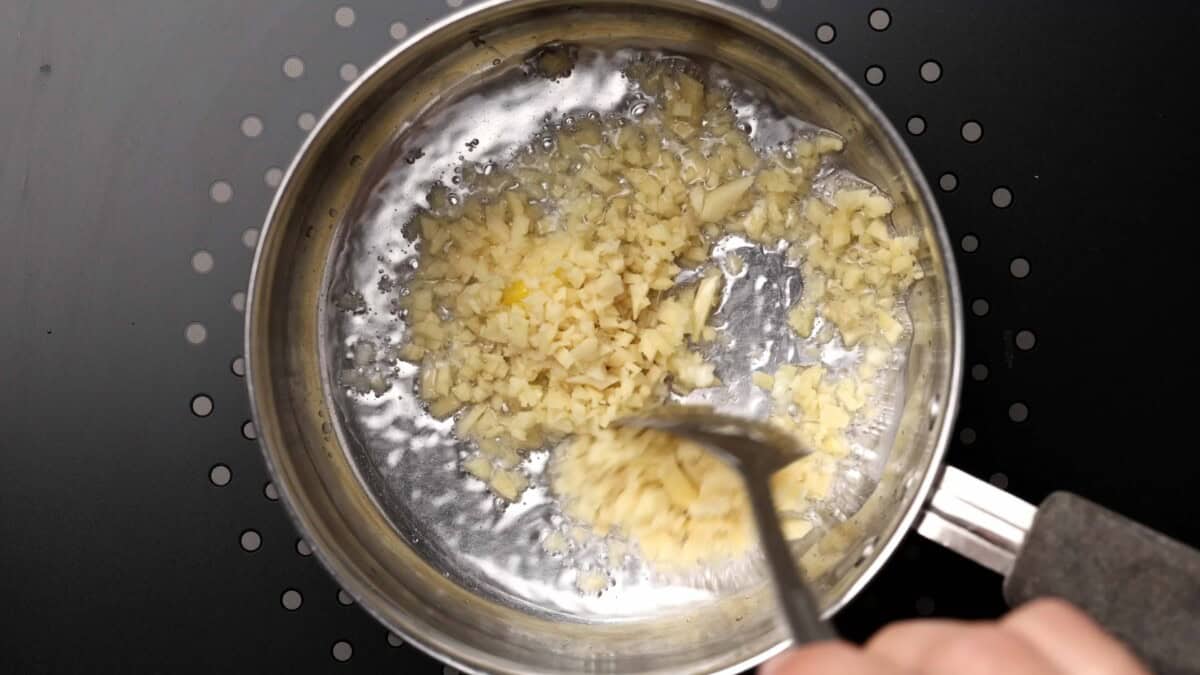 Chopped garlic and ginger being sautéed in a stainless steel pan on a stovetop, with a utensil stirring the garlic.