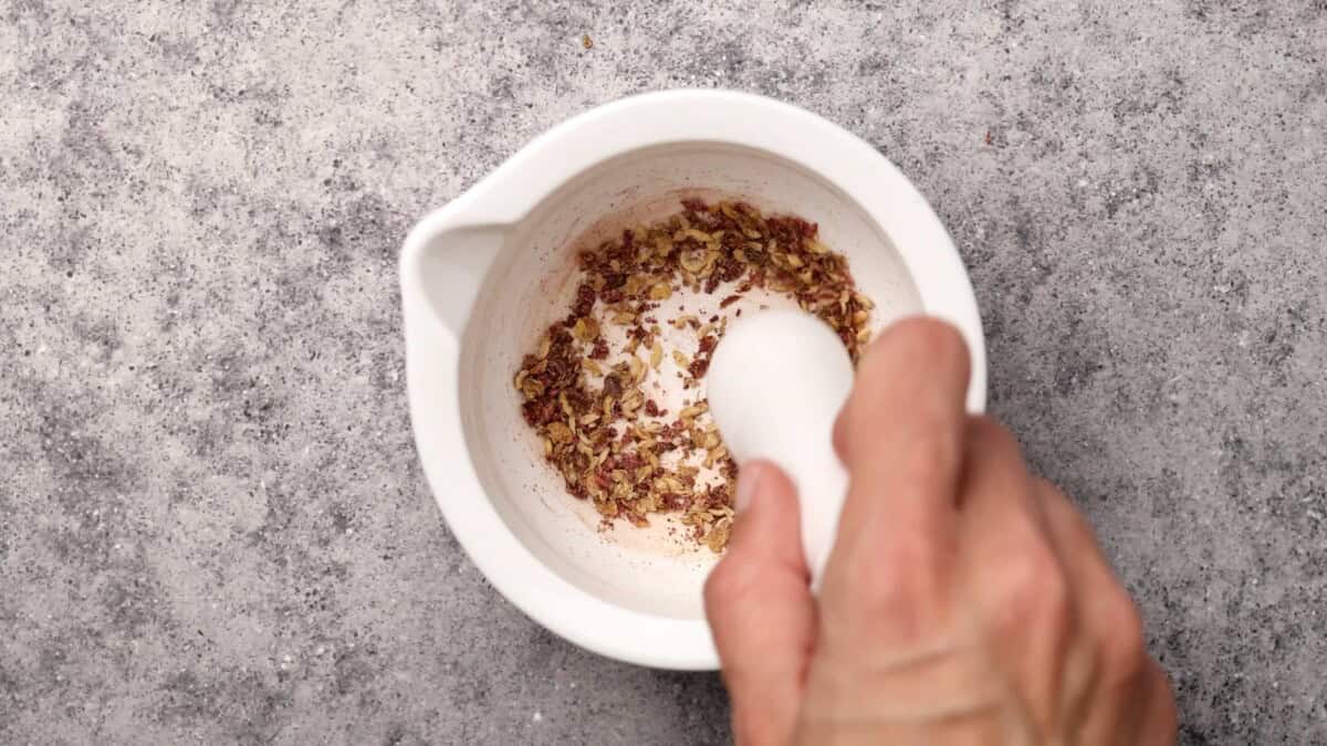 A hand uses a white mortar and pestle to grind szechuan peppercorns on a gray textured surface.