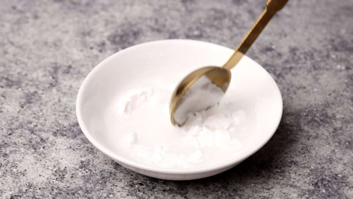 A gold spoon stirring cornstarch, with water in a small white bowl on a speckled gray surface.