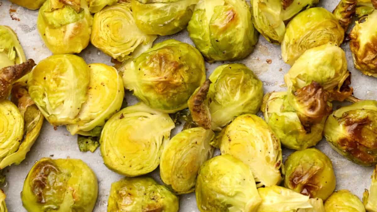Close-up of roasted Brussels sprouts, halved and spread out on a baking sheet. The sprouts have golden-brown, crispy edges and a tender, slightly glossy appearance.