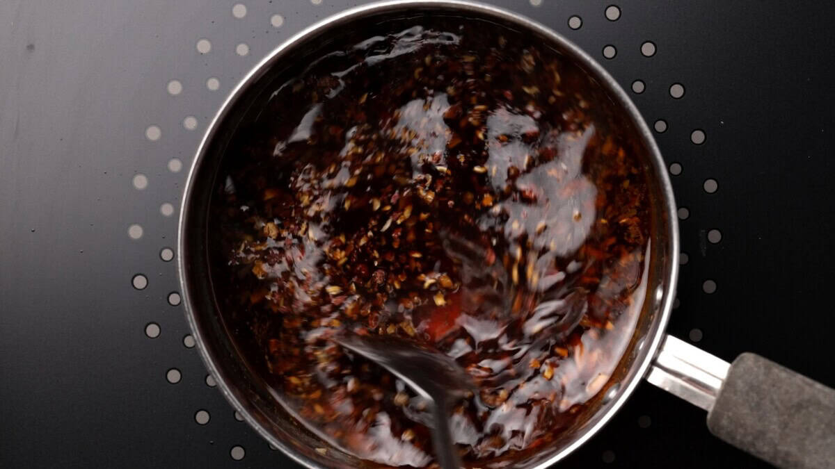 A top-down view of a pot on a black stovetop, filled with kung pao sauce being stirred with a spoon.