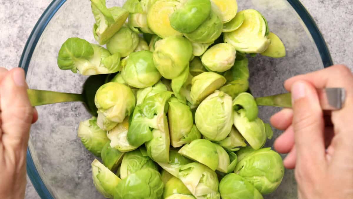 A bowl filled with halved Brussels sprouts being tossed with serving utensils, viewed from above on a gray surface.