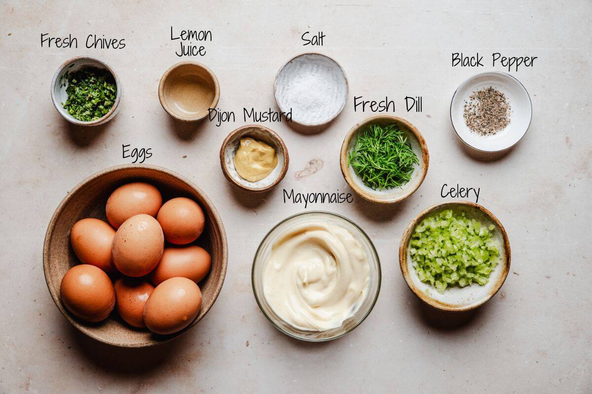 A top-down view of ingredients in bowls for egg salad: fresh chives, lemon juice, salt, black pepper, fresh dill, eggs, Dijon mustard, mayonnaise, and chopped celery, all labeled on a light surface.