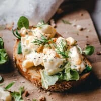 An open-faced sandwich topped with creamy egg salad, fresh greens, and dill sits on a wooden board, surrounded by scattered herbs and crumbs. Text above and below promotes "Pinch and Swirl: The BEST Egg Salad.