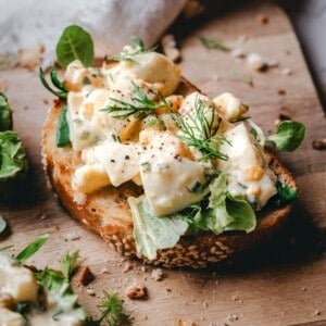 A slice of seeded bread topped with creamy egg salad, fresh greens, and garnished with dill, on a wooden surface with scattered herbs and crumbs.