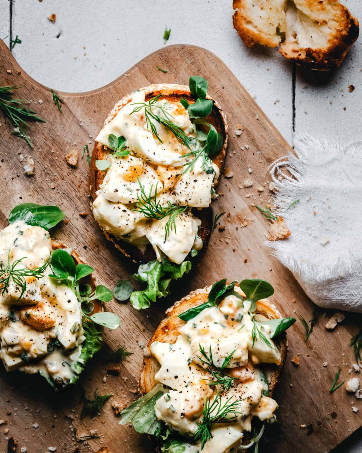 Three slices of toasted bread topped with creamy egg salad, fresh greens, and dill are arranged on a wooden board with scattered crumbs and a piece of torn bread nearby.