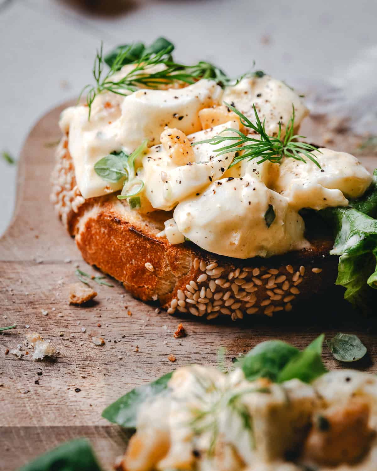 A slice of sesame bread topped with creamy egg salad, fresh greens, and dill, garnished with cracked black pepper, on a wooden board.