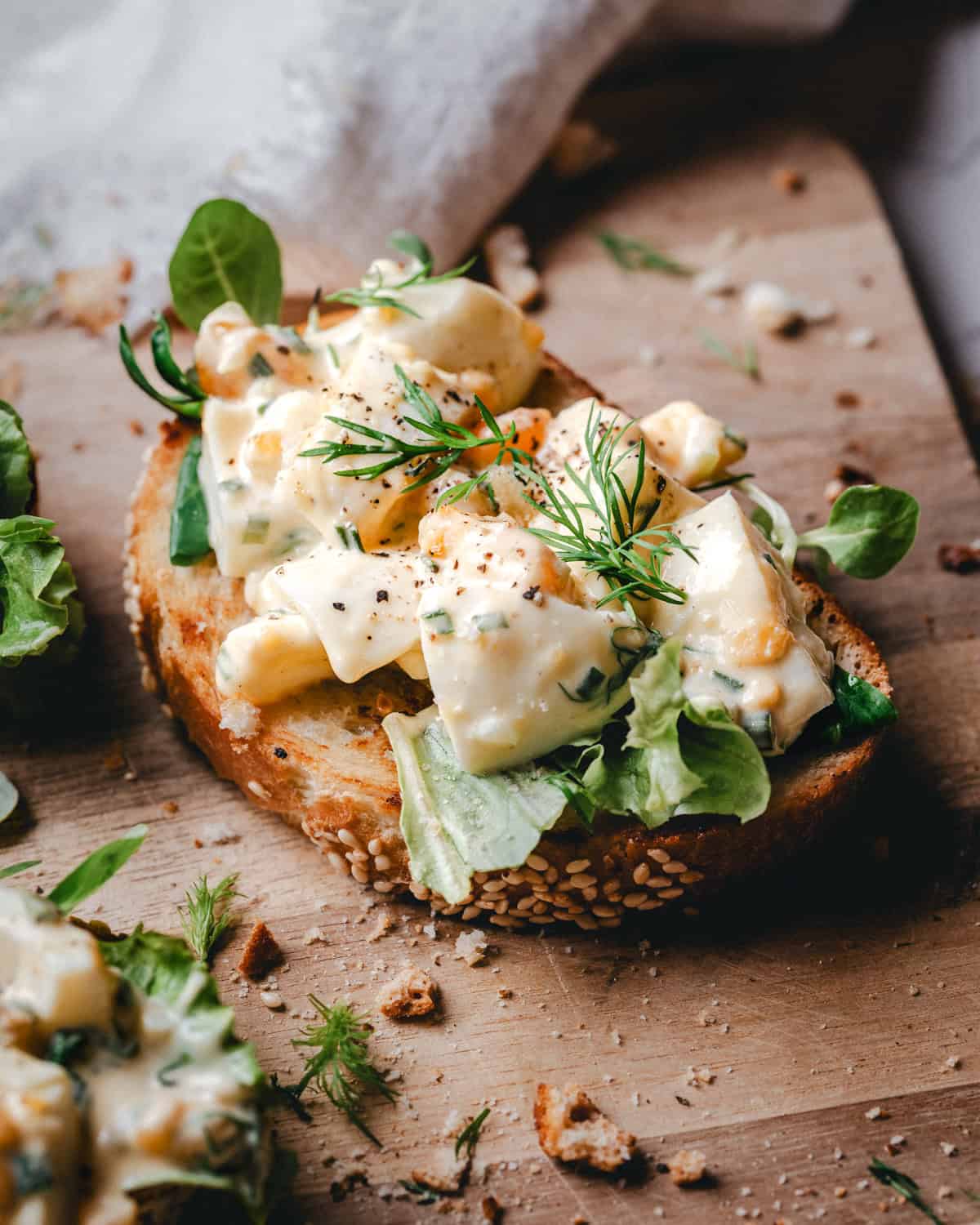 A slice of seeded bread topped with leafy greens, creamy egg salad, fresh dill, and cracked black pepper, resting on a wooden board with scattered crumbs and herbs.
