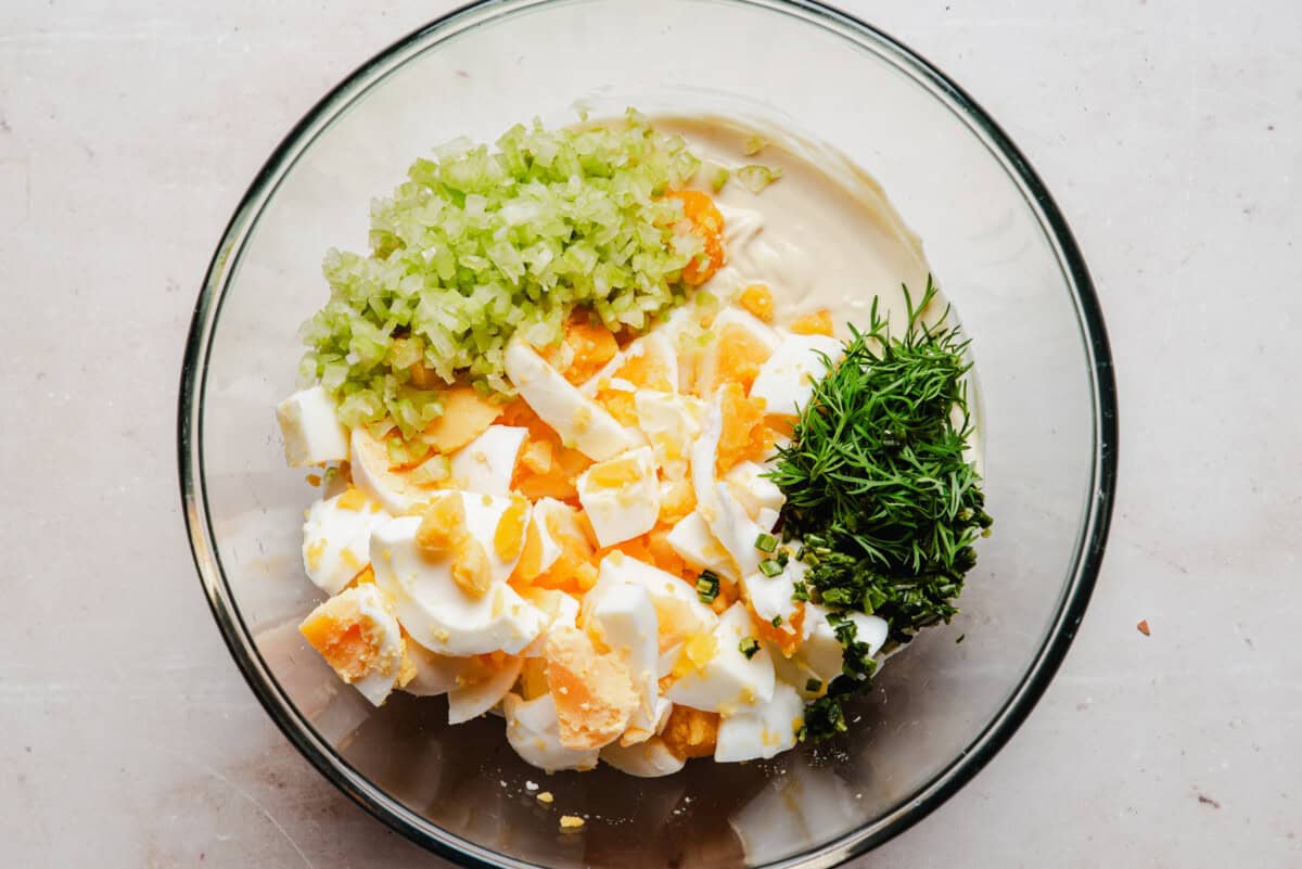A glass bowl containing chopped hard-boiled eggs, diced celery, fresh dill, and mayonnaise on a light surface, ready to be mixed for egg salad.