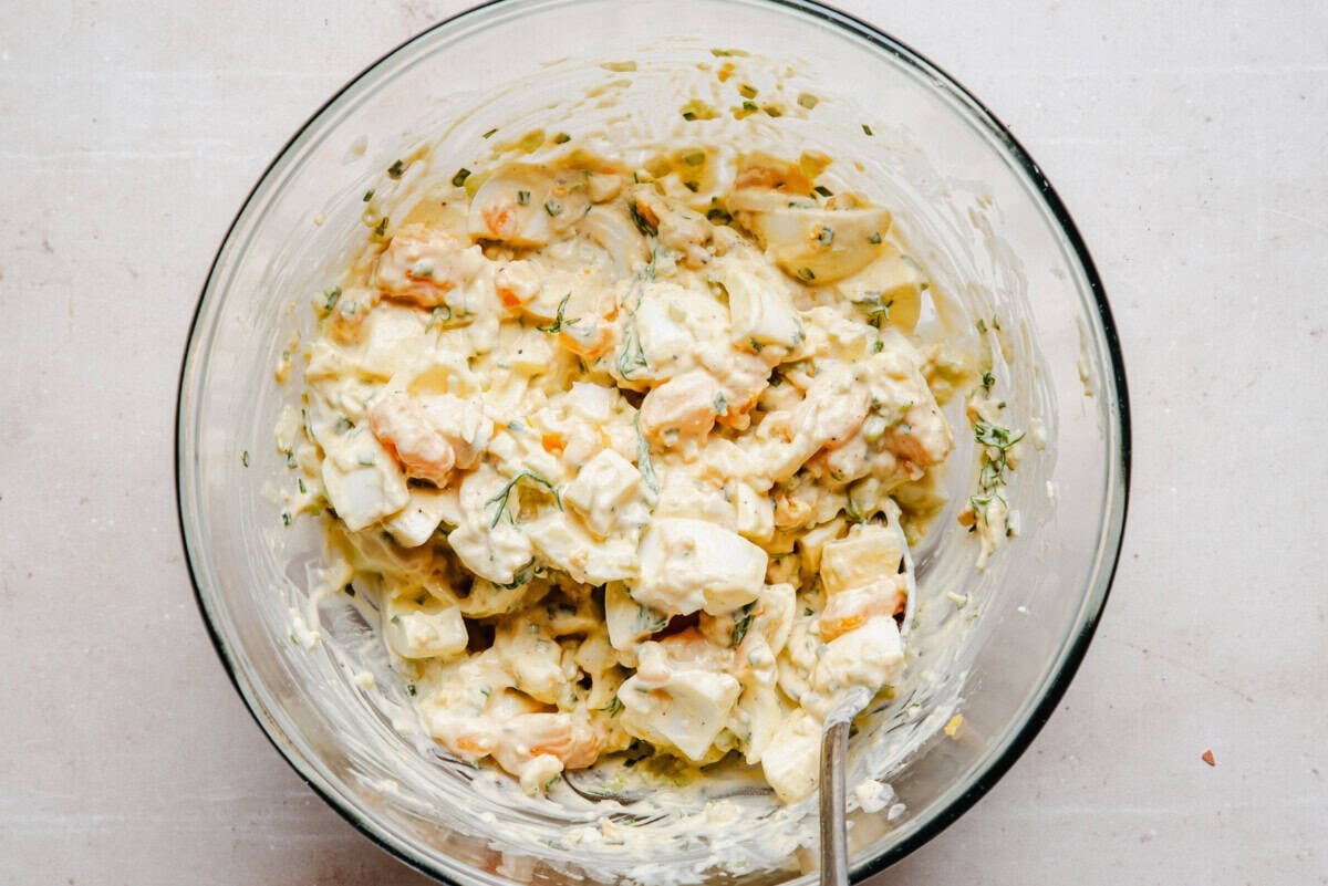 A glass bowl filled with creamy egg salad, featuring chunks of hard-boiled eggs, mayonnaise, herbs, and diced vegetables, with a spoon resting inside.