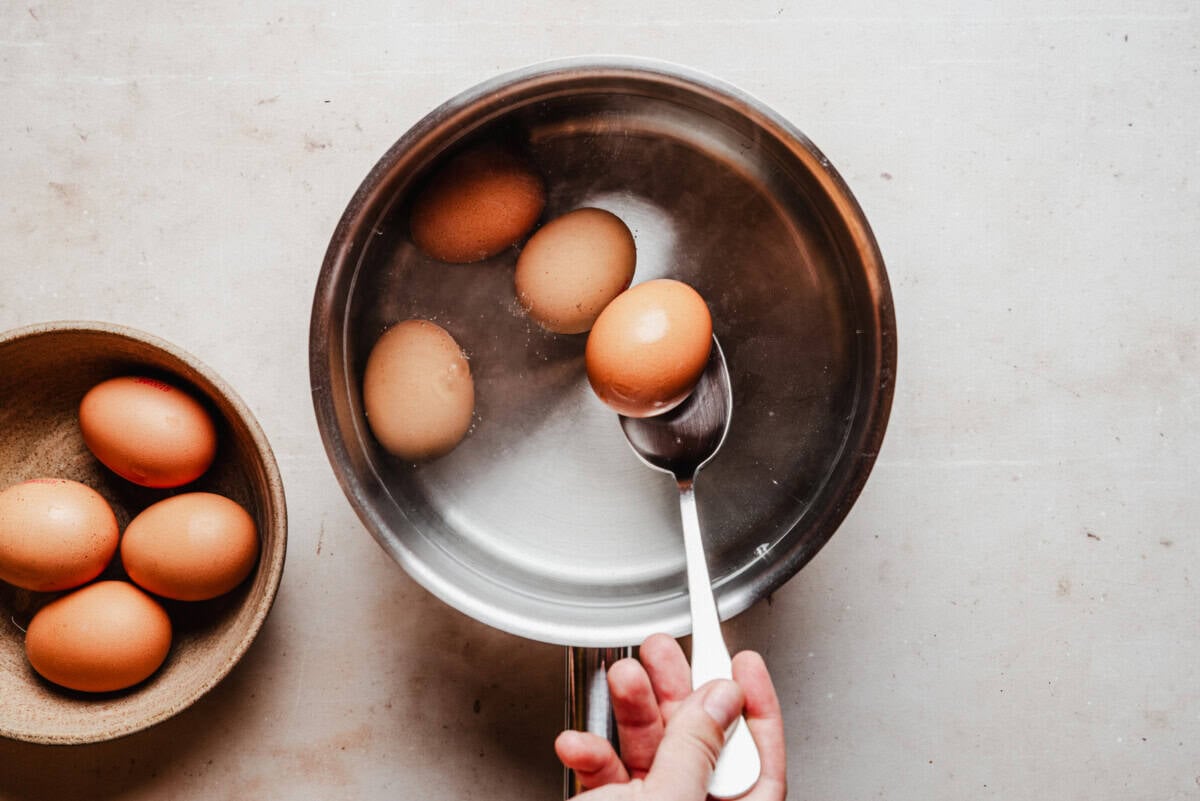 A hand uses a spoon to place a brown egg into a pot of water, while a bowl with more brown eggs sits nearby on a light countertop.