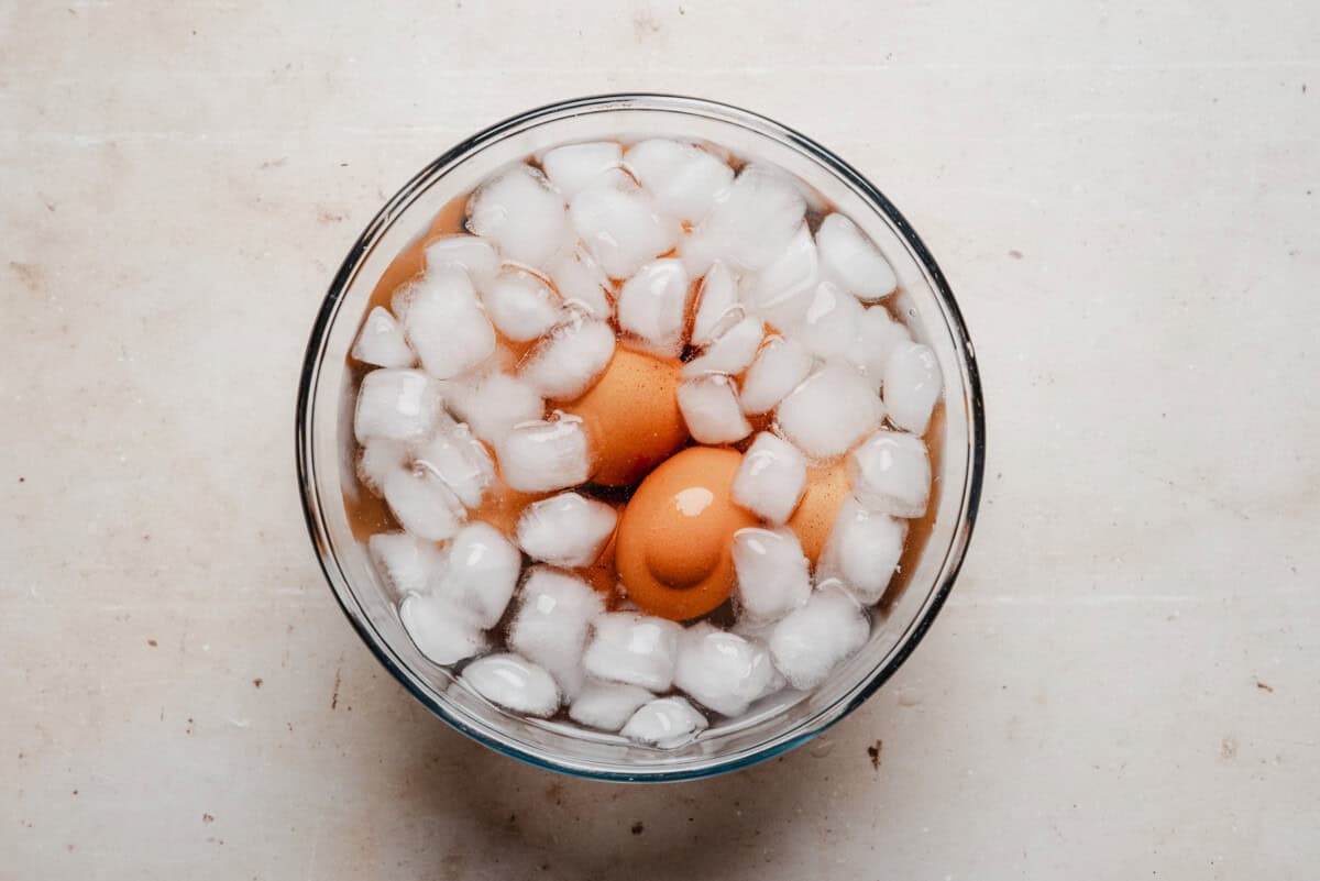 A glass bowl filled with ice cubes and water contains two brown eggs, seen from above on a light-colored surface.