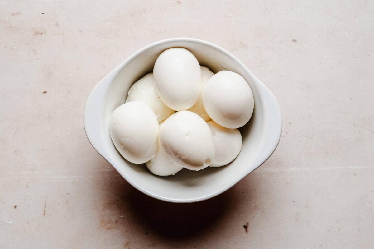 A white bowl filled with seven peeled hard-boiled eggs sits on a light-colored, textured surface.