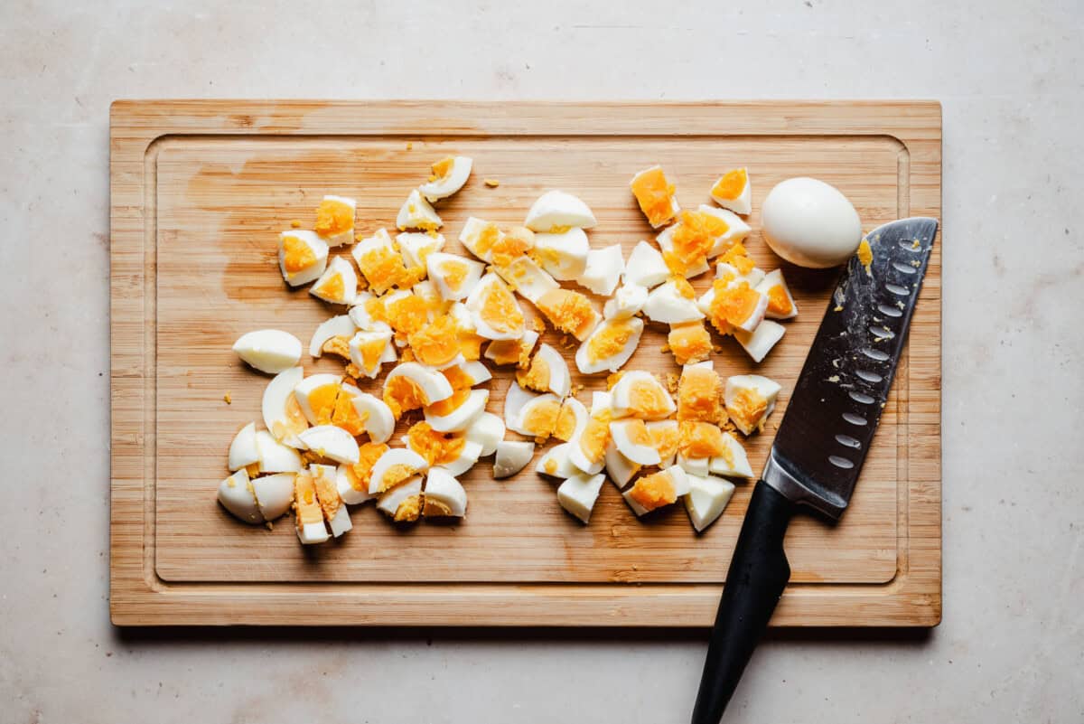 Chopped hard-boiled eggs scattered on a wooden cutting board, with a kitchen knife placed to the right and an uncut egg nearby.