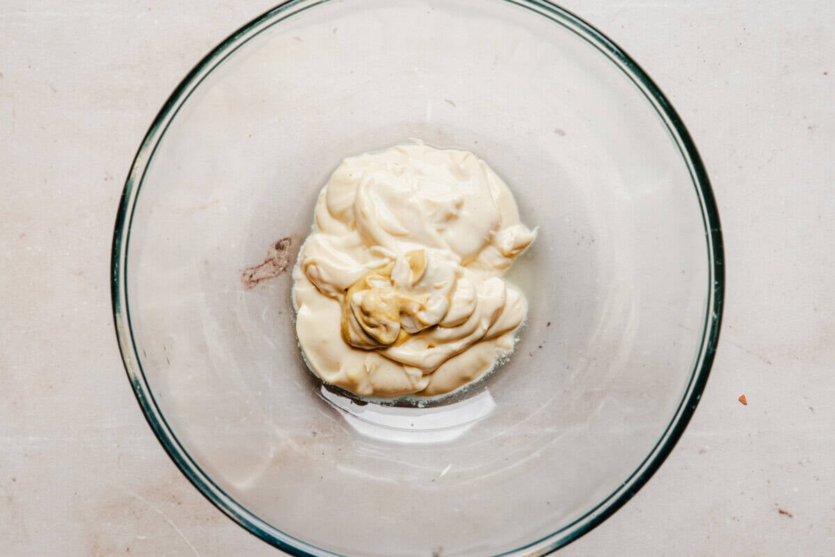 A glass bowl containing a dollop of mayonnaise and a small amount of mustard on a light countertop.