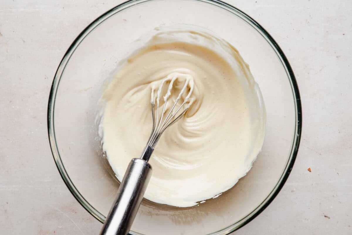 A metal whisk rests in a glass bowl filled with creamy, smooth batter or sauce on a light-colored surface.