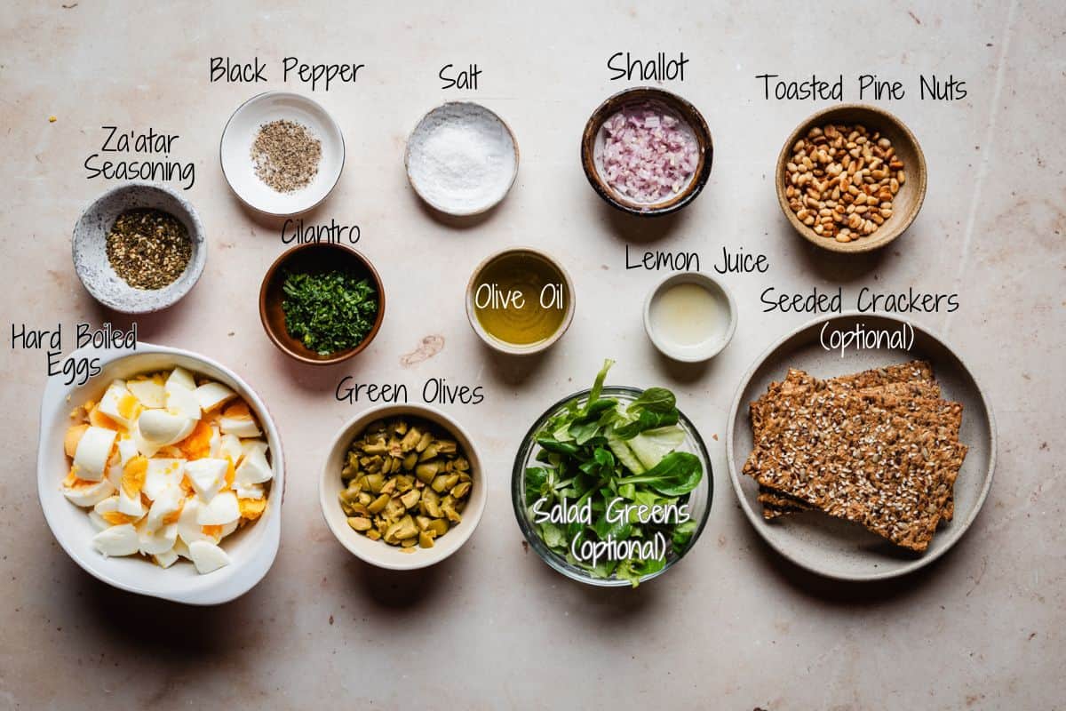 A top view of bowls and plates containing black pepper, salt, shallot, toasted pine nuts, za’atar, cilantro, olive oil, lemon juice, hard-boiled eggs, green olives, salad greens, and seeded crackers. Labels identify each item.