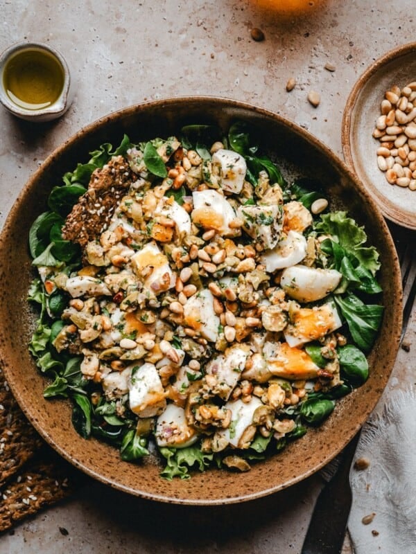 A bowl of leafy green salad topped with Mediterranean Egg Salad. Surrounding the bowl are crackers, a small bowl of pine nuts, a cup of olive oil, and a white napkin on a beige surface.