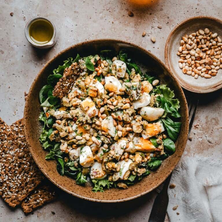 A bowl of leafy green salad topped with Mediterranean Egg Salad. Surrounding the bowl are crackers, a small bowl of pine nuts, a cup of olive oil, and a white napkin on a beige surface.