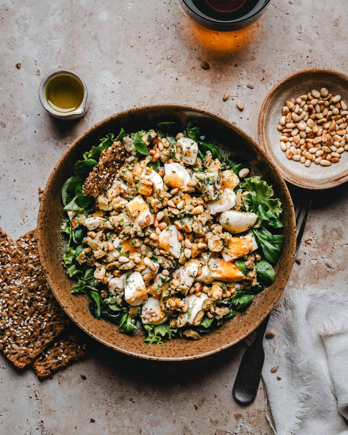 A bowl of fresh Mediterranean Egg Salad topped with sliced egg, cheese, herbs, and pine nuts, surrounded by a cup of oil, a bowl of pine nuts, seed crackers, a fork, and a white napkin on a light stone surface.