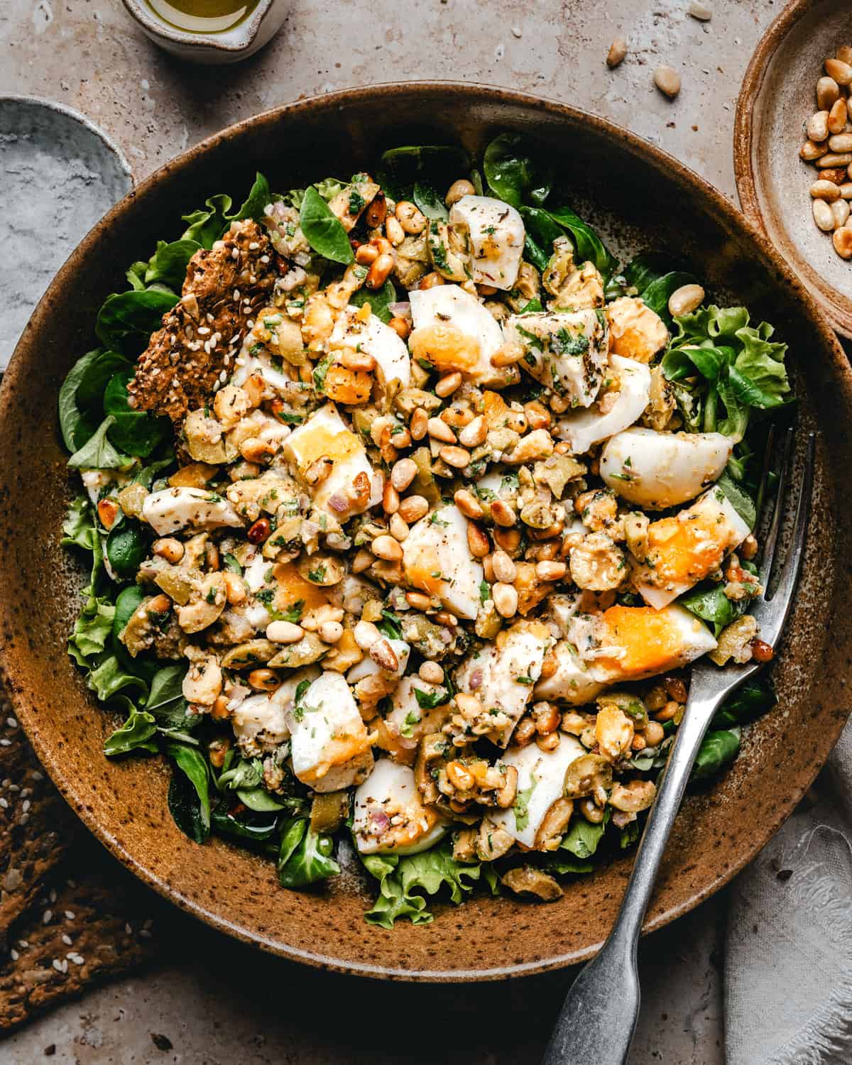 A bowl of Mediterranean Egg Salad with leafy greens, sliced boiled eggs, chopped nuts or seeds, and a creamy dressing, served with a fork and crispbread on the side.