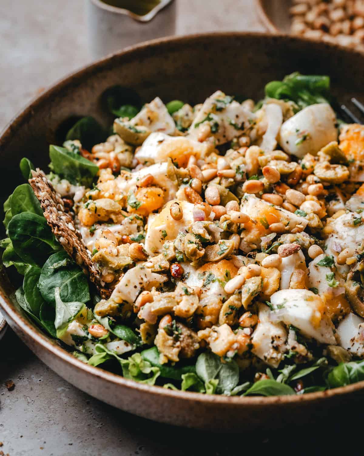 A close-up of a Mediterranean Egg Salad in a brown bowl topped with chopped boiled eggs, pine nuts, seeds, greens, and herbs, with a crisp cracker on the side. A fork and a small dish of dressing are nearby.