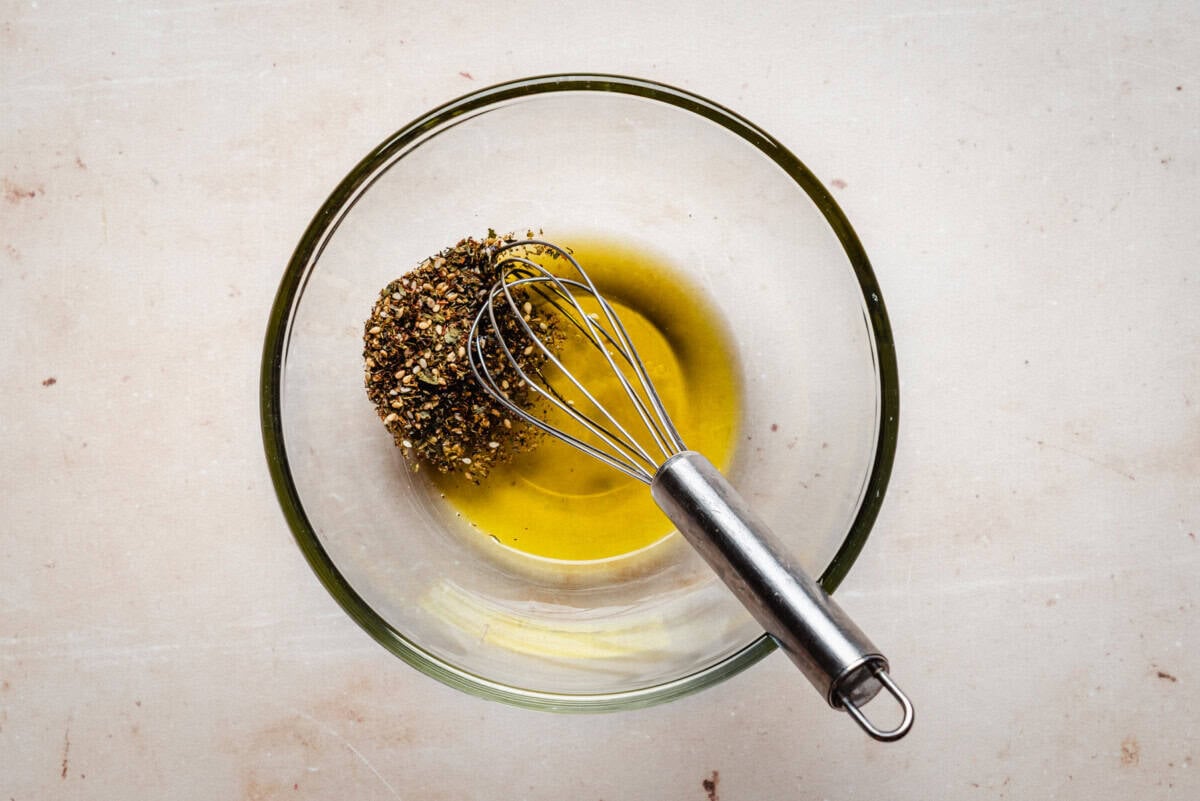 A clear glass bowl containing olive oil and za'atar seasoning sits on a light surface. A metal whisk rests inside the bowl, ready to mix the ingredients.