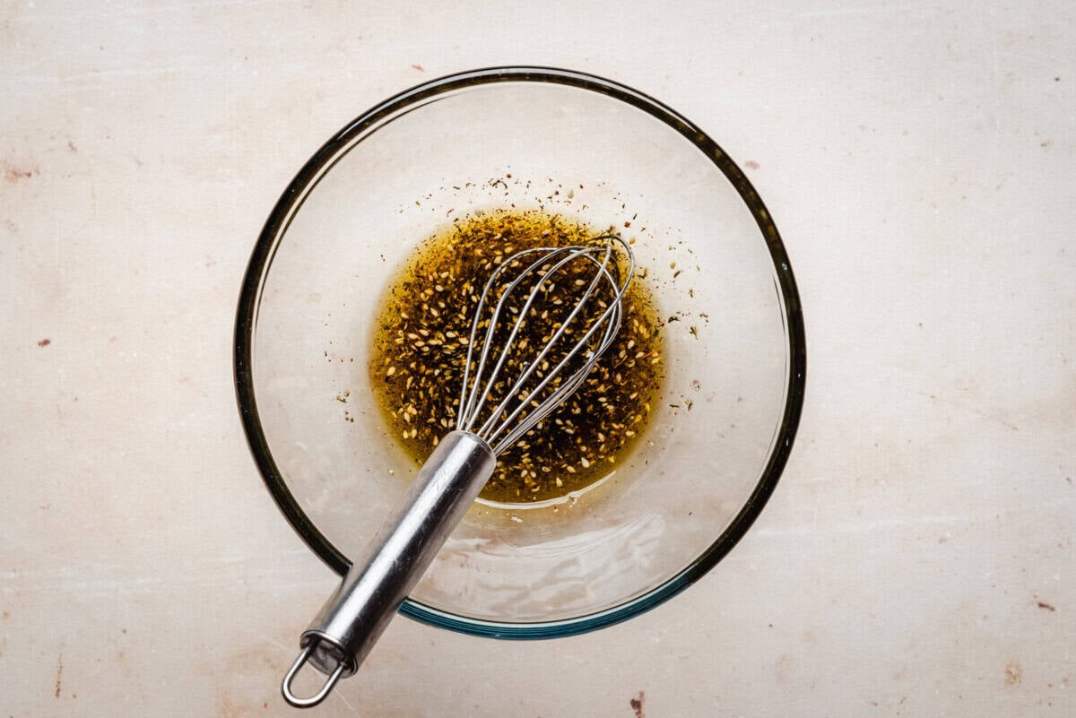 A metal whisk rests in a glass bowl containing a mixture of oil, vinegar, and cracked black pepper on a light-colored countertop.