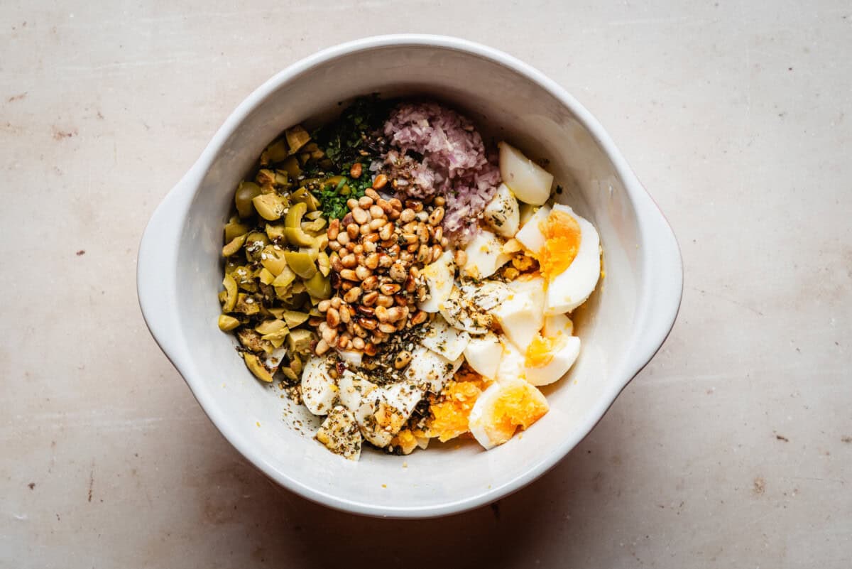 A white bowl contains chopped hard-boiled eggs, diced green olives, pine nuts, chopped red onion, and herbs, all arranged separately on a light countertop.